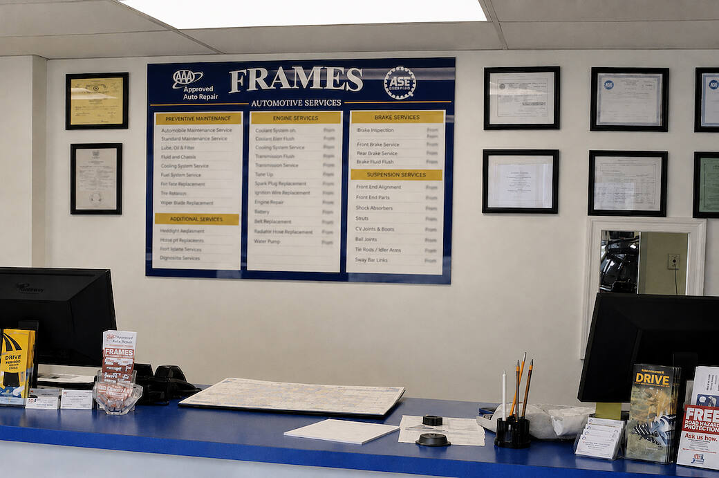 The board behind the counter listing the shop guarantee and a menu of priced services, under the AAA and ASE marks, with framed certificates on the wall beside it
